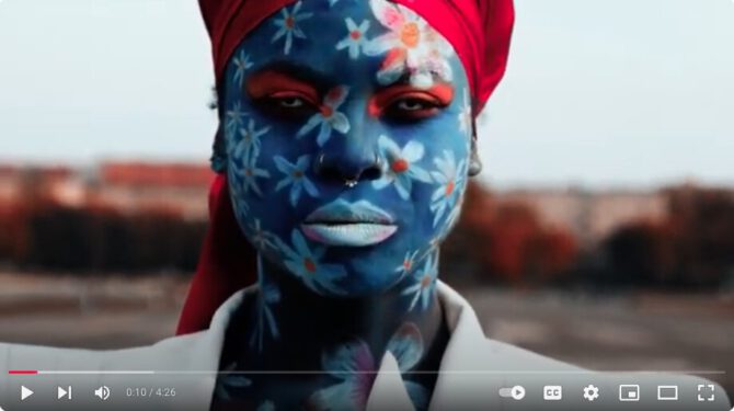 GodXXX Noirphiles wears facepaint of blue daisies on a blue background, a white suit jacket and a bright read head wrap