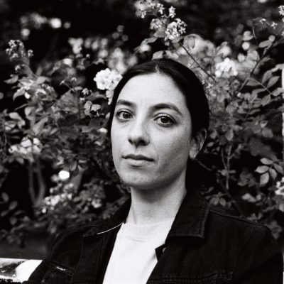 black and white image of terri looking at the camera, hair tied back, in front of a bush of flowers