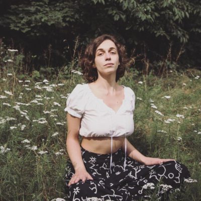 Ioana looks at the camera wearing a cropped white blouse and sits yoga style in a field